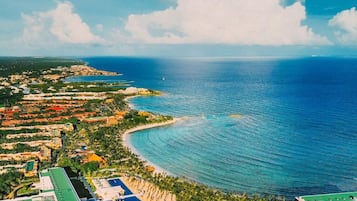 Vista aérea de un complejo turístico costero con un edificio grande y curvo de techo verde, rodeado de abundante vegetación y un mar azul cristalino.