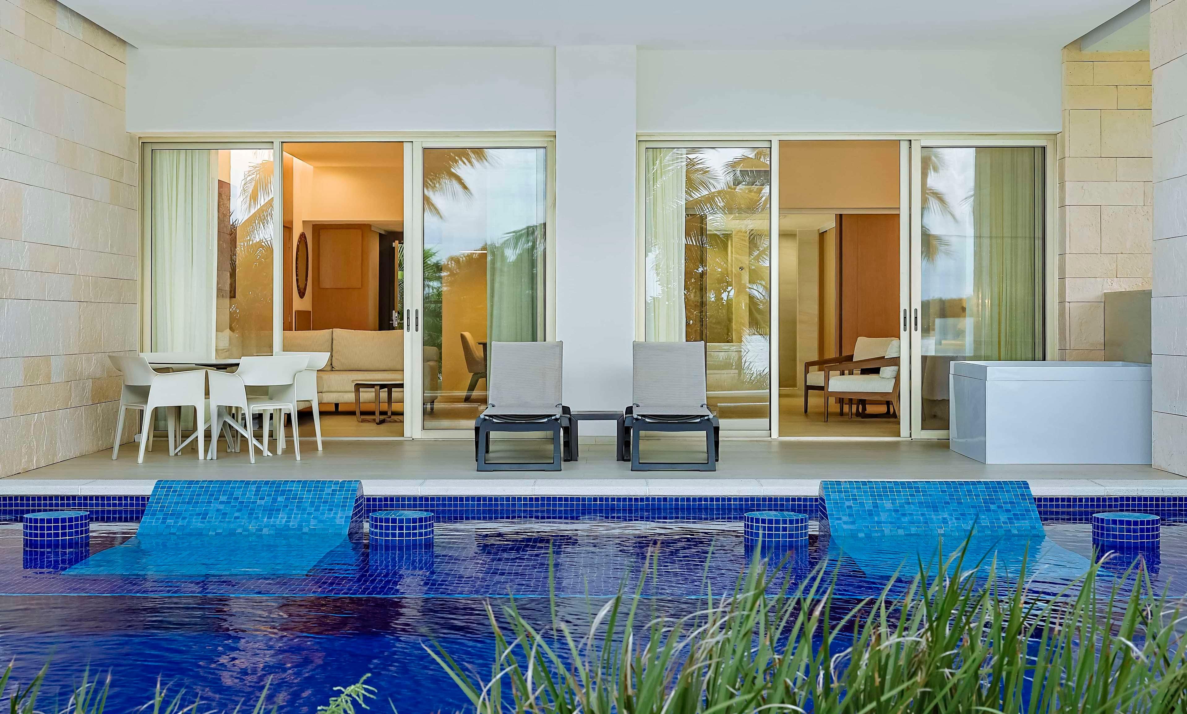 A modern outdoor pool area with a glass balustrade, two lounge chairs, and a dining table with chairs.