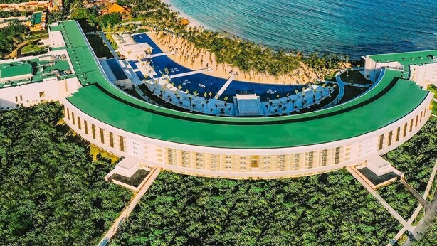 Vista aerea di una località balneare con un ampio edificio curvo dotato di tetto verde, immerso nel verde e con un mare cristallino.