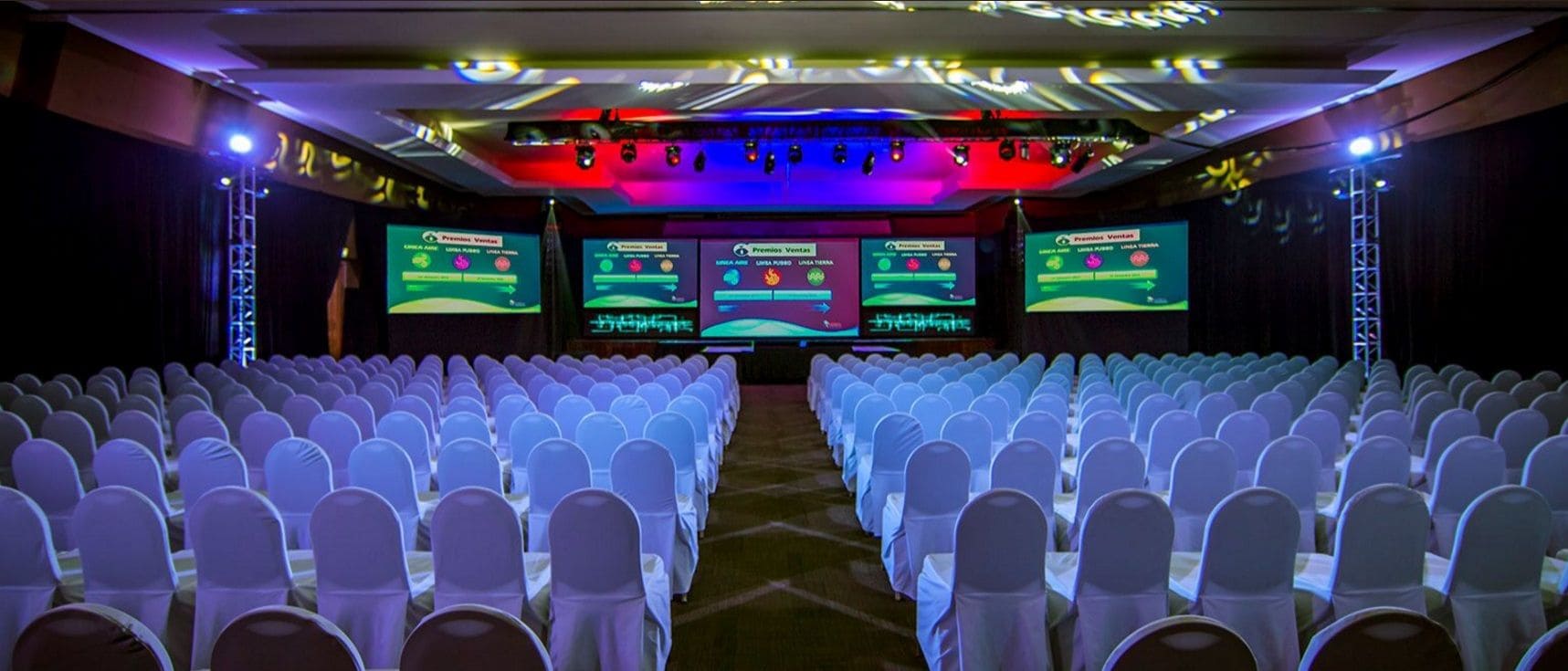 A conference room with rows of white chairs, a stage with a large screen, and a podium.