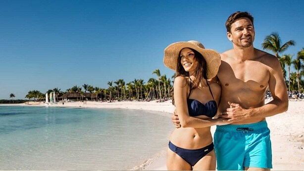 Une scène de plage avec une eau d’un bleu limpide, des palmiers et un couple qui s’embrasse.