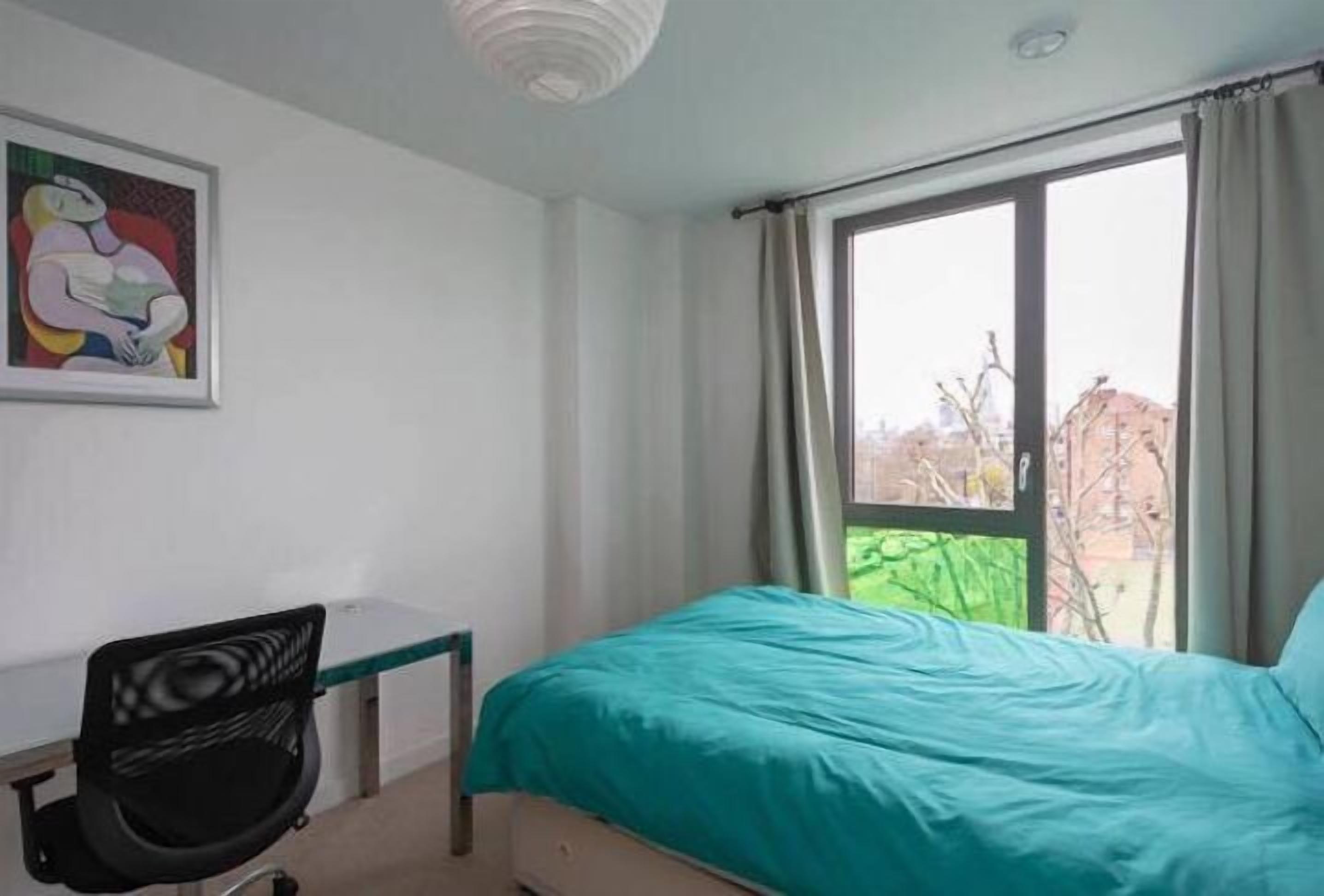 A bedroom with a bed, a desk, and a chair. There is a window with curtains, and a painting on the wall.