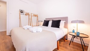 A neatly made bed with white linens, a round side table with a lamp, and a wooden floor.