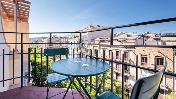 A balcony with a table and chairs, offering a view of a cityscape with buildings and greenery.