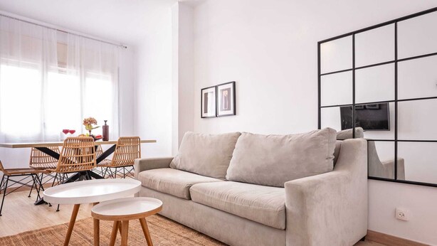 A modern living room with a beige sofa, a small round table, wicker chairs, and a large window with sheer curtains. - Sweett | Rocafort 604 (Barcelona)
