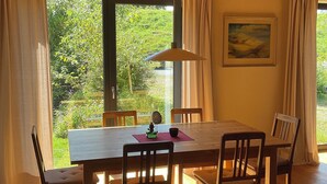 A dining area with a wooden table and chairs, large windows with curtains, and a painting on the wall. - Vacation home at Süderdeich in Friedrichskoog (Friedrichskoog)