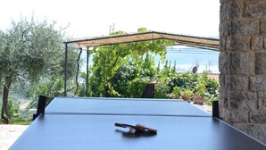 Mesa de ping pong al aire libre con red, rodeada de muros de piedra y follaje.