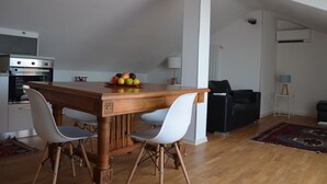 Una cocina moderna con una mesa de madera, sillas blancas y un frutero. Hay un sofá negro y una lámpara en la sala.