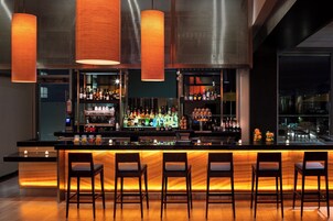 A modern bar with a long counter, multiple shelves of bottles, and three bar stools.
