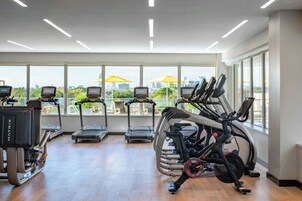 Academia moderna com esteiras, elípticos e bicicletas ergométricas. Grandes janelas proporcionam vista para a paisagem urbana.