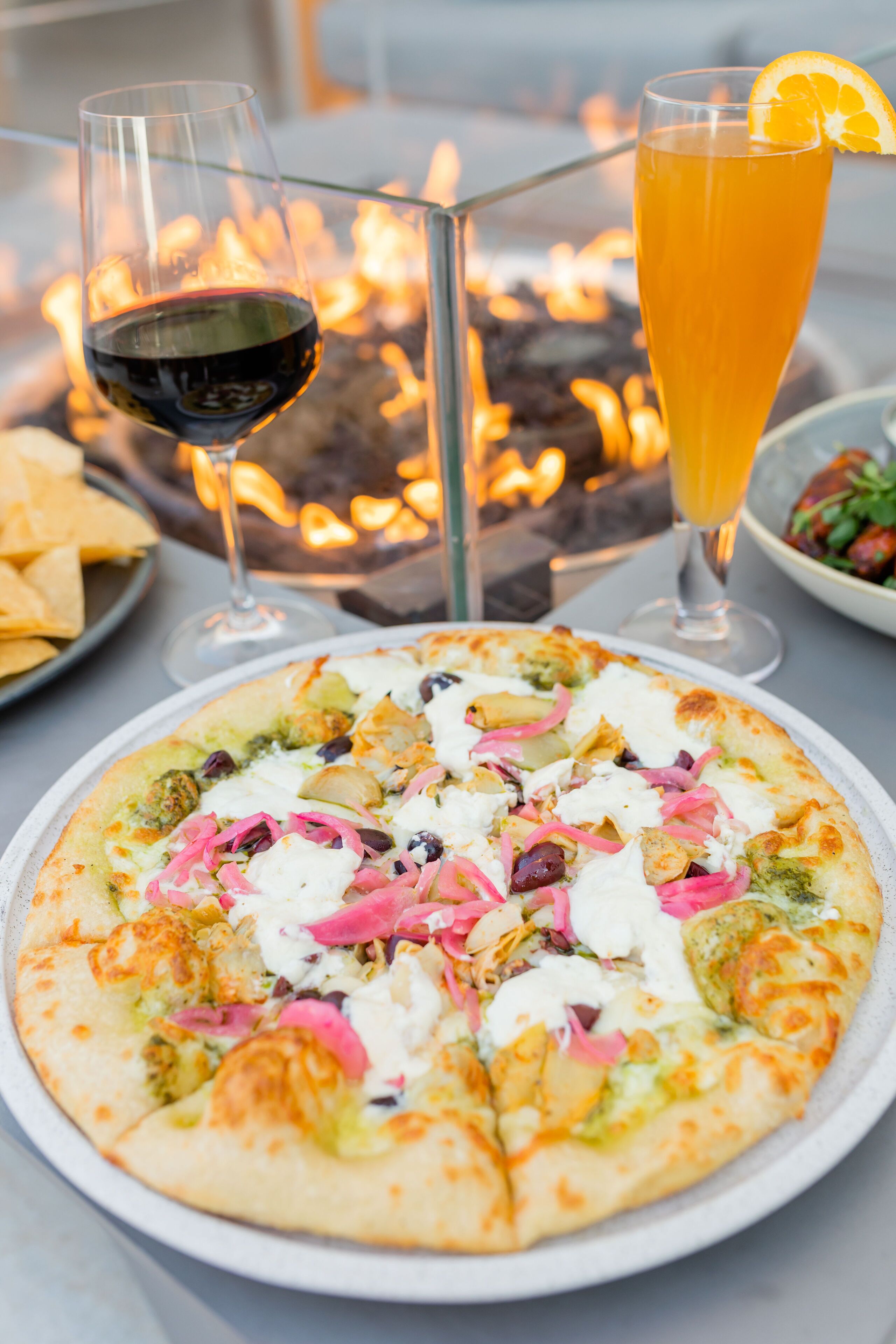 A pizza with various toppings, a glass of red wine, and a glass of orange juice on a table with a fire in the background.