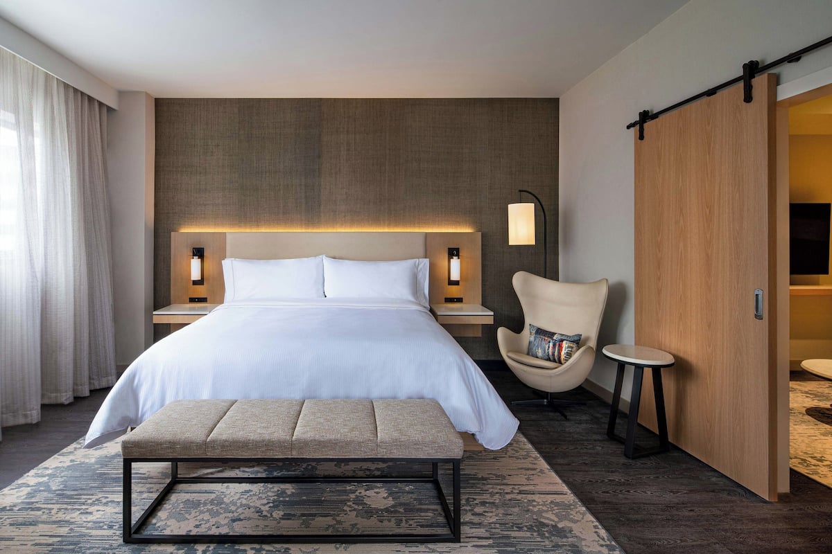 A modern hotel room with a large bed, bedside lamps, a bench, a chair, and a wooden wardrobe.