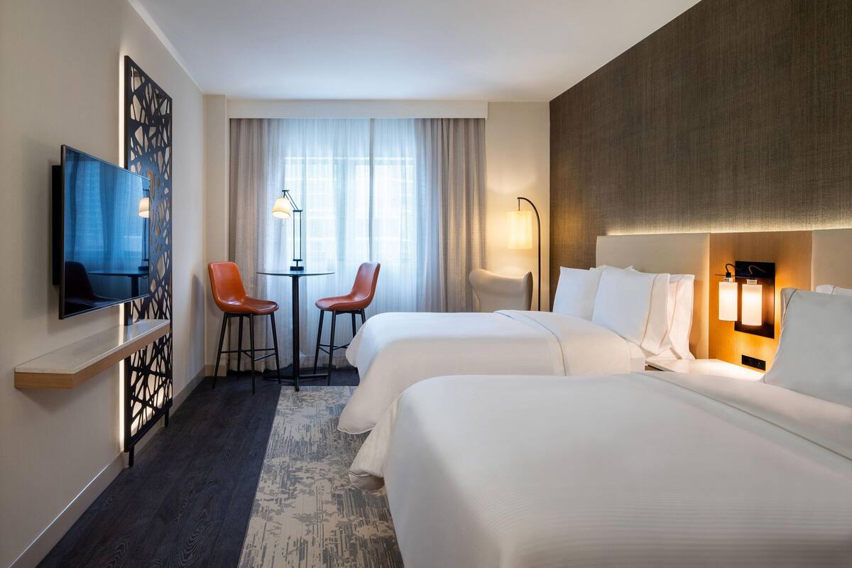 A hotel room with two beds, a TV, a desk with two chairs, a window with curtains, and a wall-mounted lamp.