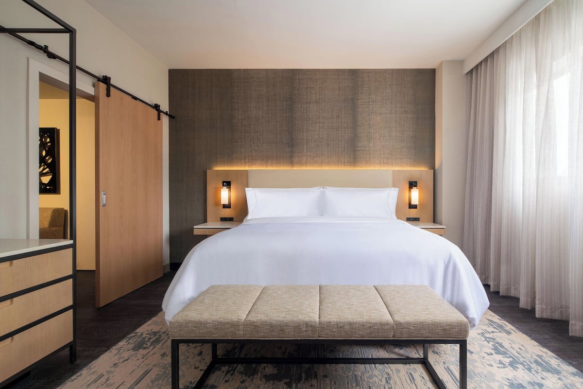 A hotel room with a large bed, bedside lamps, a bench, and a view of the outside.