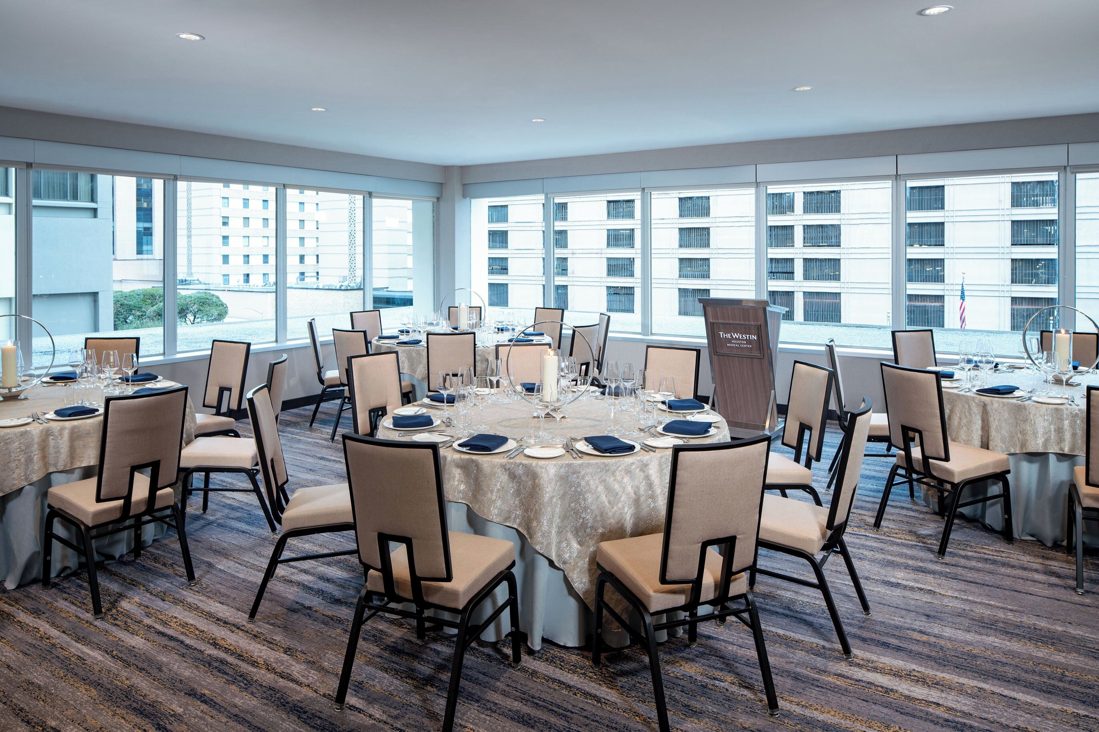 A conference room with multiple tables set for a meeting, large windows with a view of buildings, and a carpeted floor.