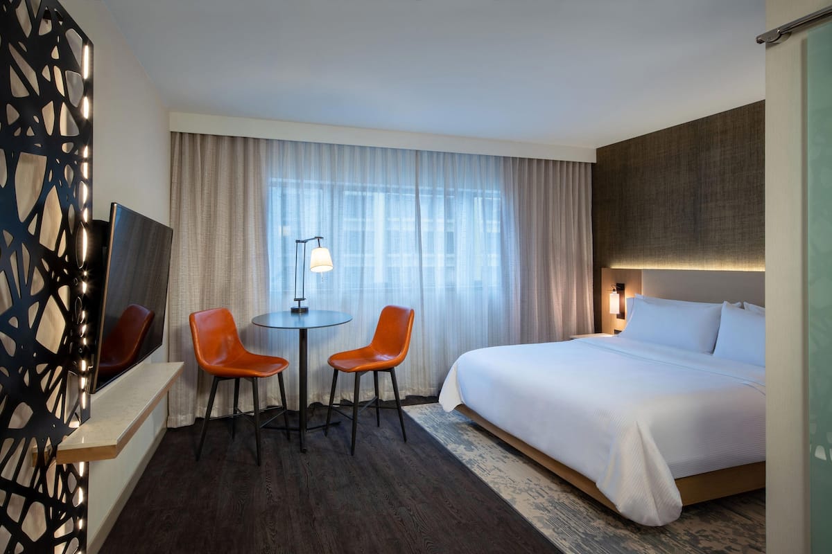 A hotel room with a bed, two orange chairs, a small table, a TV, and a window with curtains.