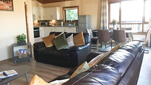 Een moderne woonkamer met een zwarte leren bank, een eethoek met stoelen, een keuken met witte kastjes en een groot raam dat zorgt voor daglicht.
