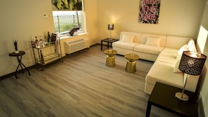 A living room with a white sofa, a glass coffee table, a side table, a lamp, a radiator, a window with a view, and a wall art piece. - Wyndham Garden North Bergen Near Secaucus (North Bergen)