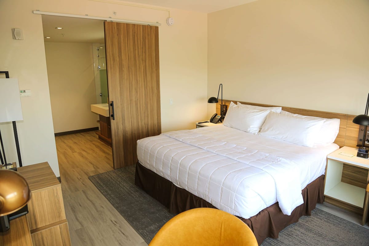 Ein Hotelzimmer mit einem großen Bett, einem Nachttisch, einer Lampe und einer Holztür zu einem Badezimmer.