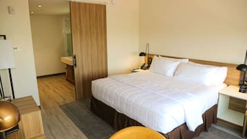 A hotel room with a large bed, bedside table, lamp, and a wooden door leading to a bathroom.