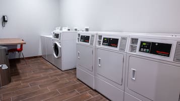 Une buanderie équipée de plusieurs machines à laver et de plusieurs sèche-linge, d’une table en bois et d’une poubelle.