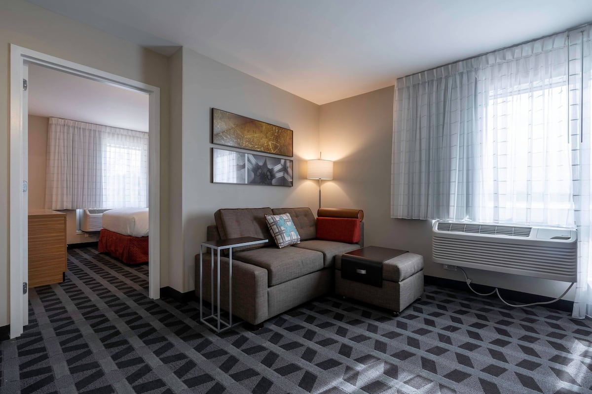A hotel room with a sofa, a small table, a lamp, a window with curtains, and an air conditioning unit.