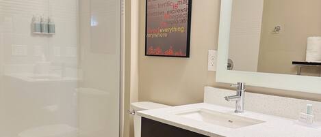 Un baño moderno con cabina de ducha de vidrio, inodoro blanco, espejo grande y tocador de madera oscura con encimera blanca.