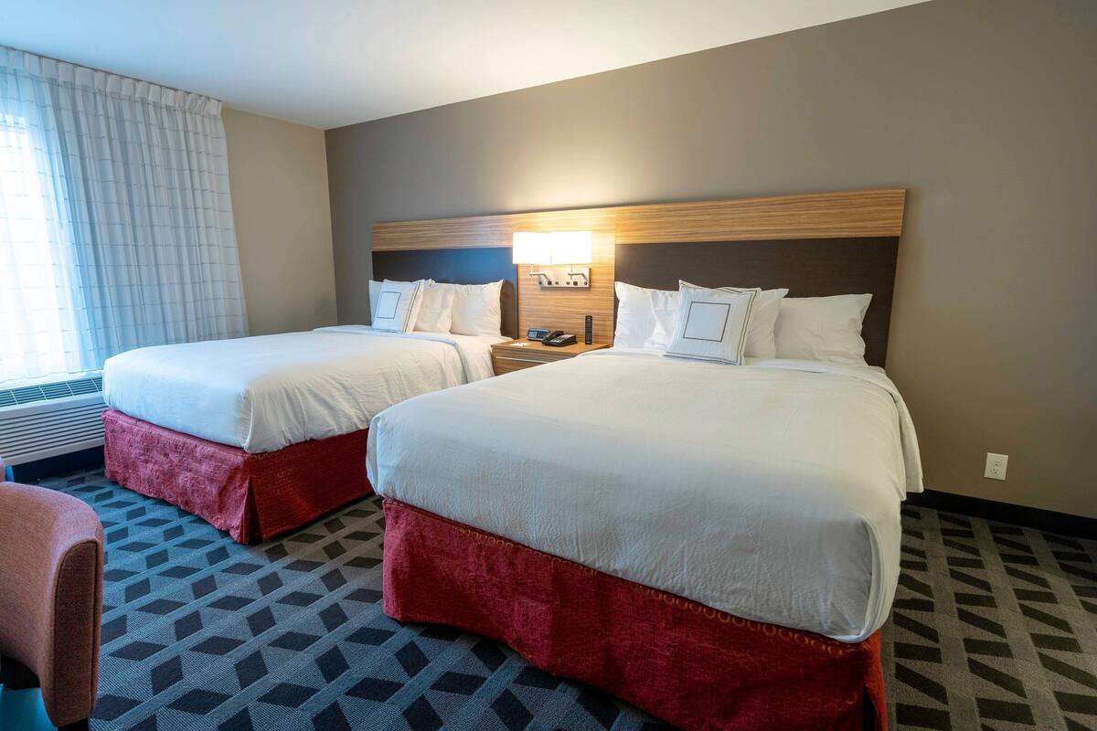 A hotel room with two beds, a nightstand with a lamp, a chair, and a patterned carpet.