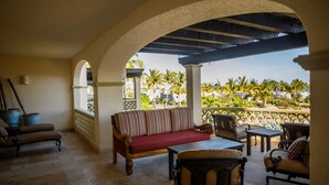 Un espacio de estar al aire libre techado con sofá y sillones, una mesa y vistas a palmeras y edificios.