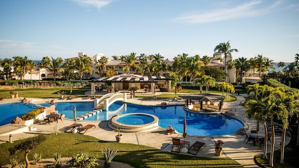 Un resort con una amplia piscina central, palmeras y un césped cuidado.