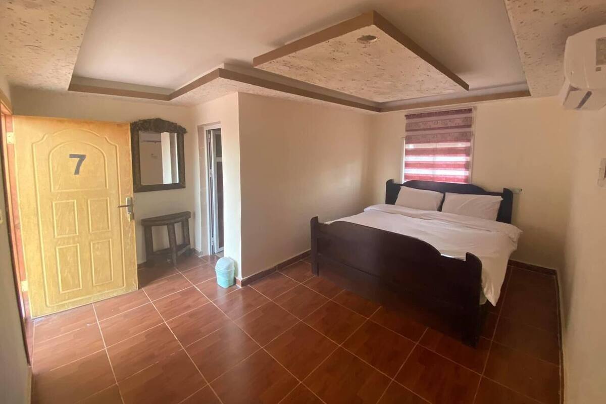 Un dormitorio con una cama, una puerta con el número 7, una mesita pequeña y un cubo de basura.