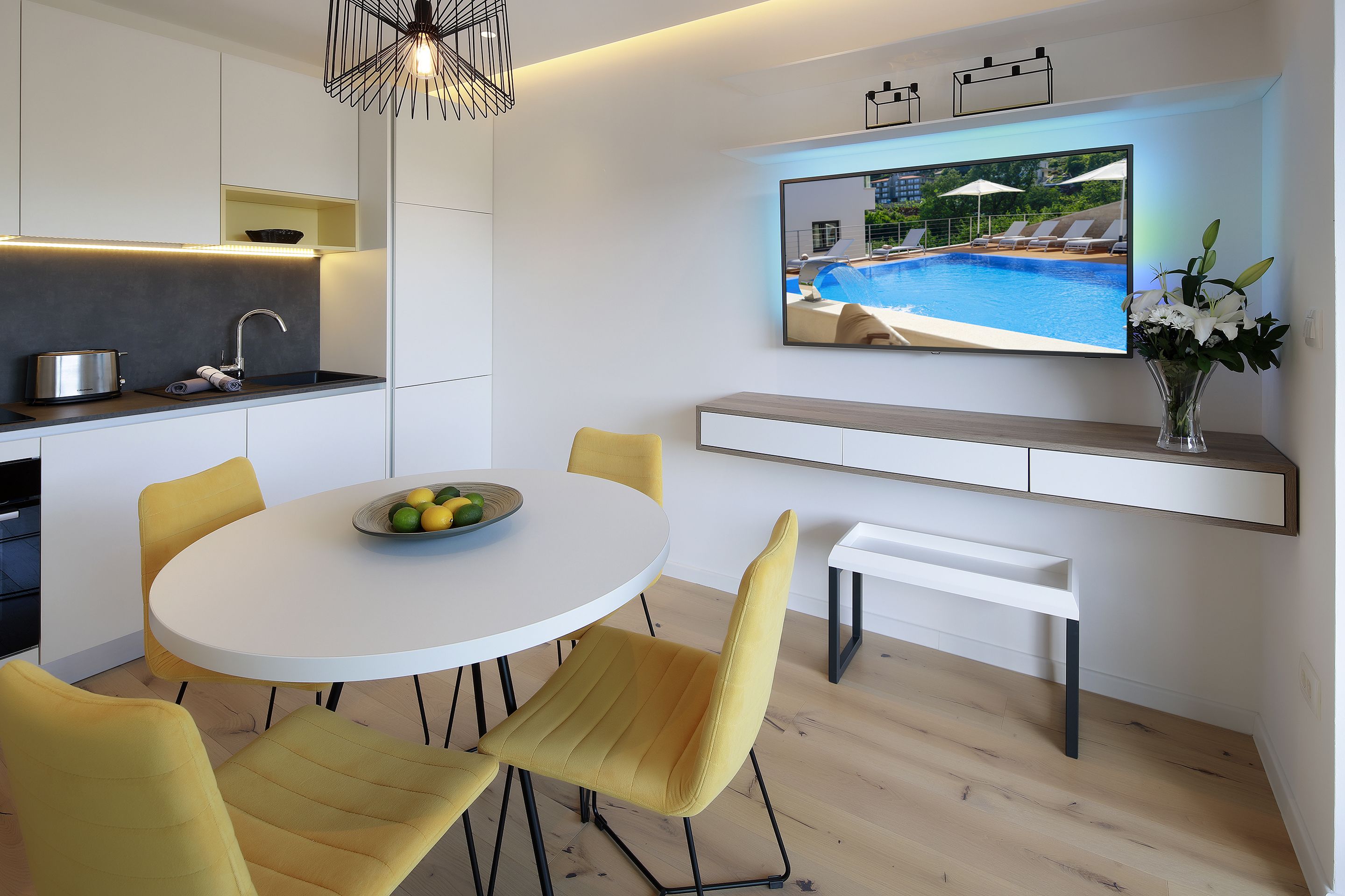 A modern kitchen with a built-in oven, a dining area with a round table and yellow chairs, and a TV mounted on the wall displaying a pool scene.