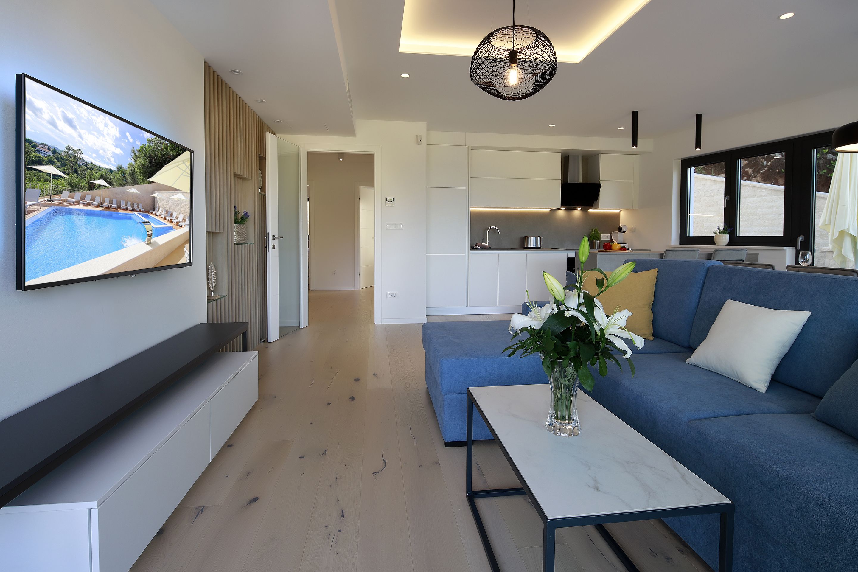 A modern living room with a large TV, a blue sofa, a white coffee table, and a kitchen area in the background.