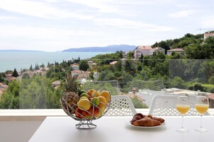 A table with a fruit bowl, croissants, and glasses of orange juice, overlooking a scenic view of a lake and houses.