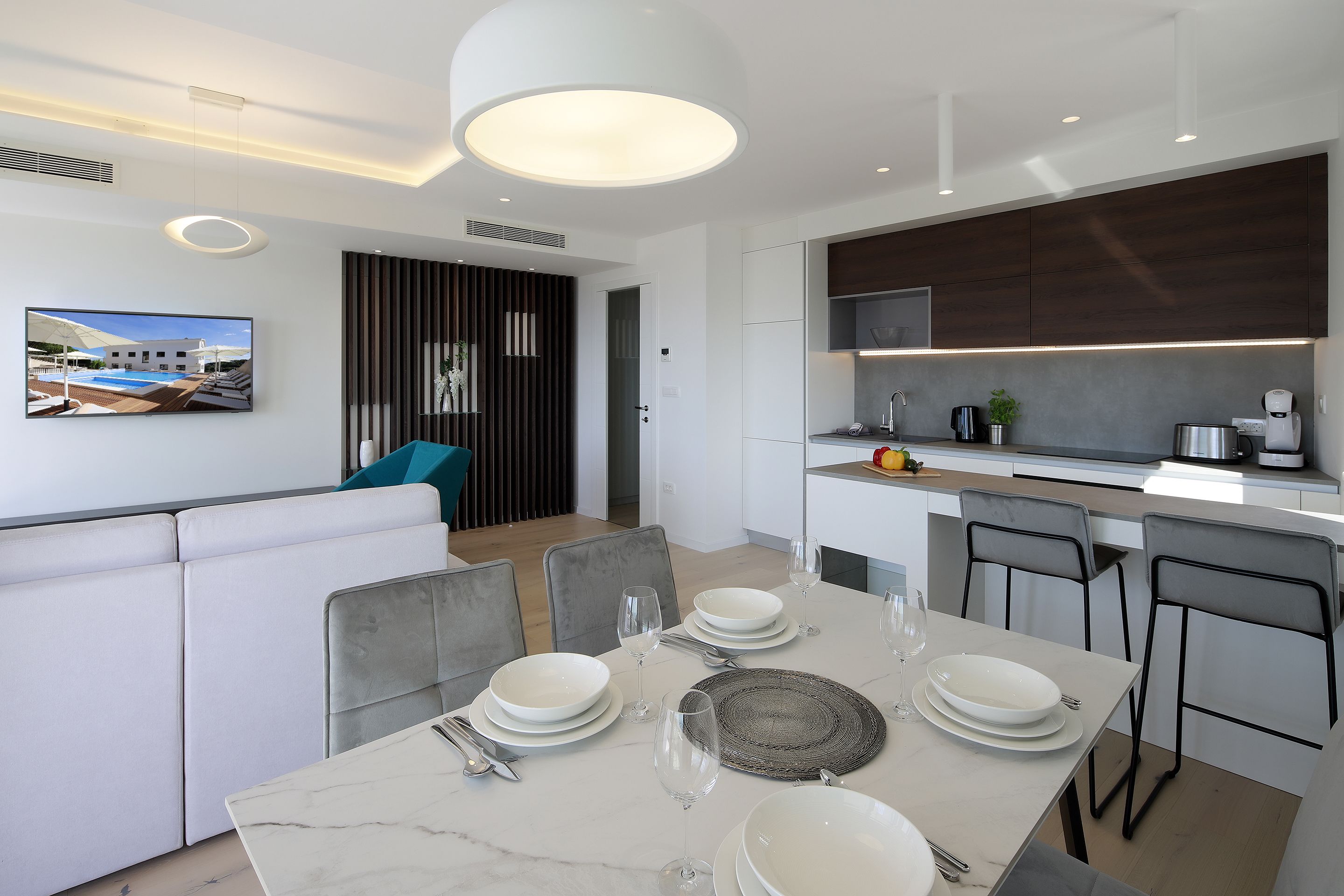 Un comedor moderno con una mesa de mármol dispuesta para cuatro personas, una cocina con armarios de madera oscura y un amplio ventanal con vistas a una piscina.