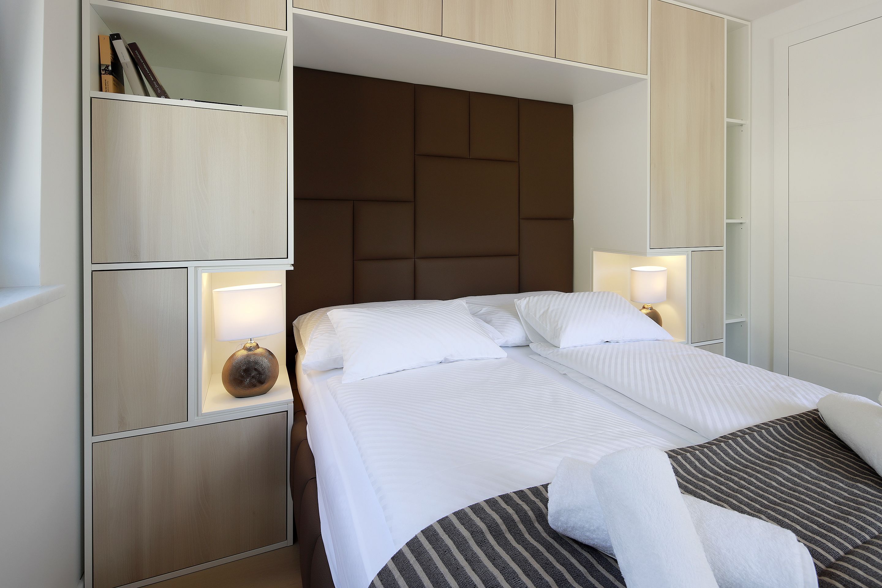 A neatly made bed with white linens and a striped blanket, a bedside lamp, and a shelf with books and a decorative item.