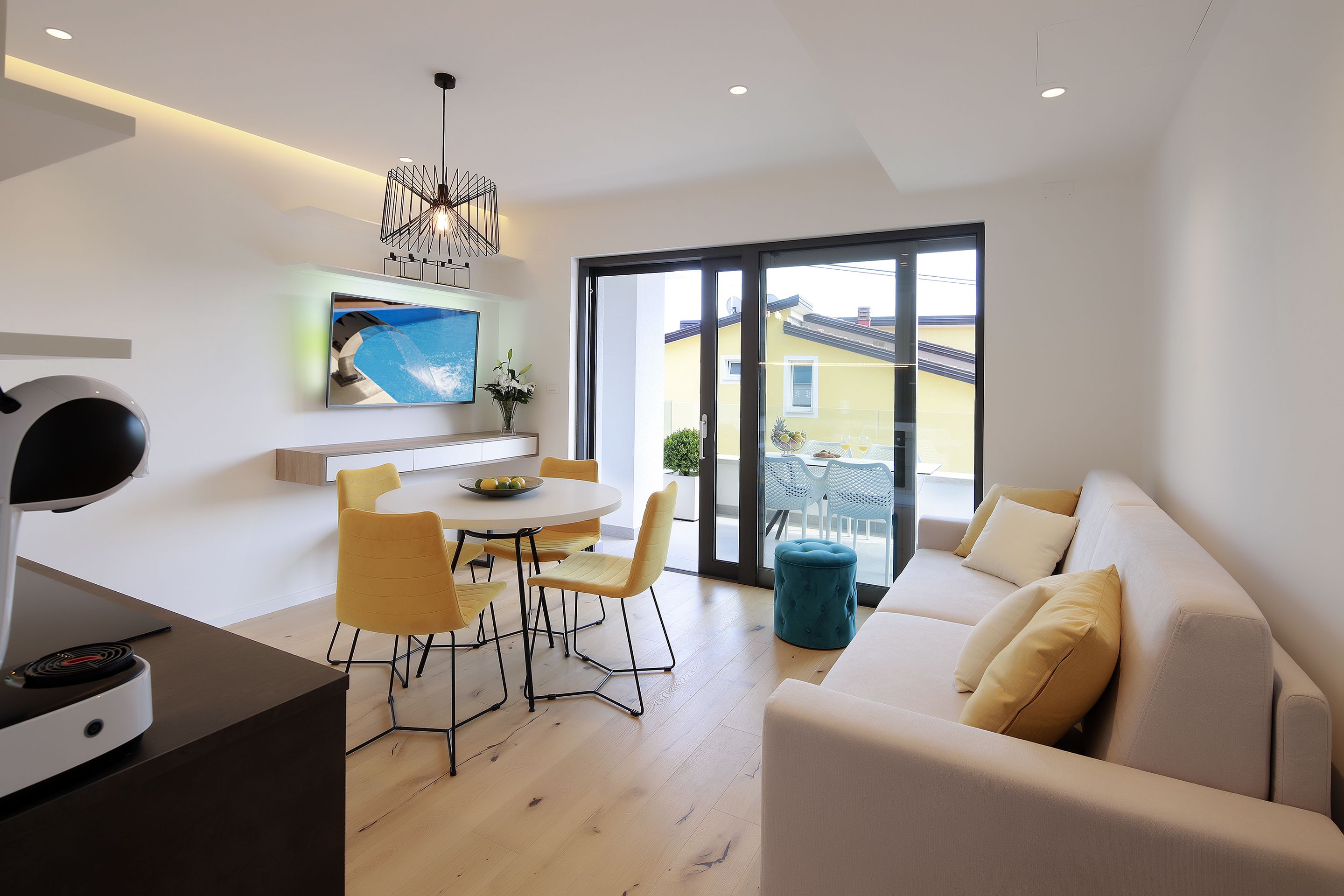Un salon moderne comprenant un canapé, une table à manger avec des chaises, un téléviseur fixé au mur et une grande porte-fenêtre coulissante donnant sur un espace extérieur.