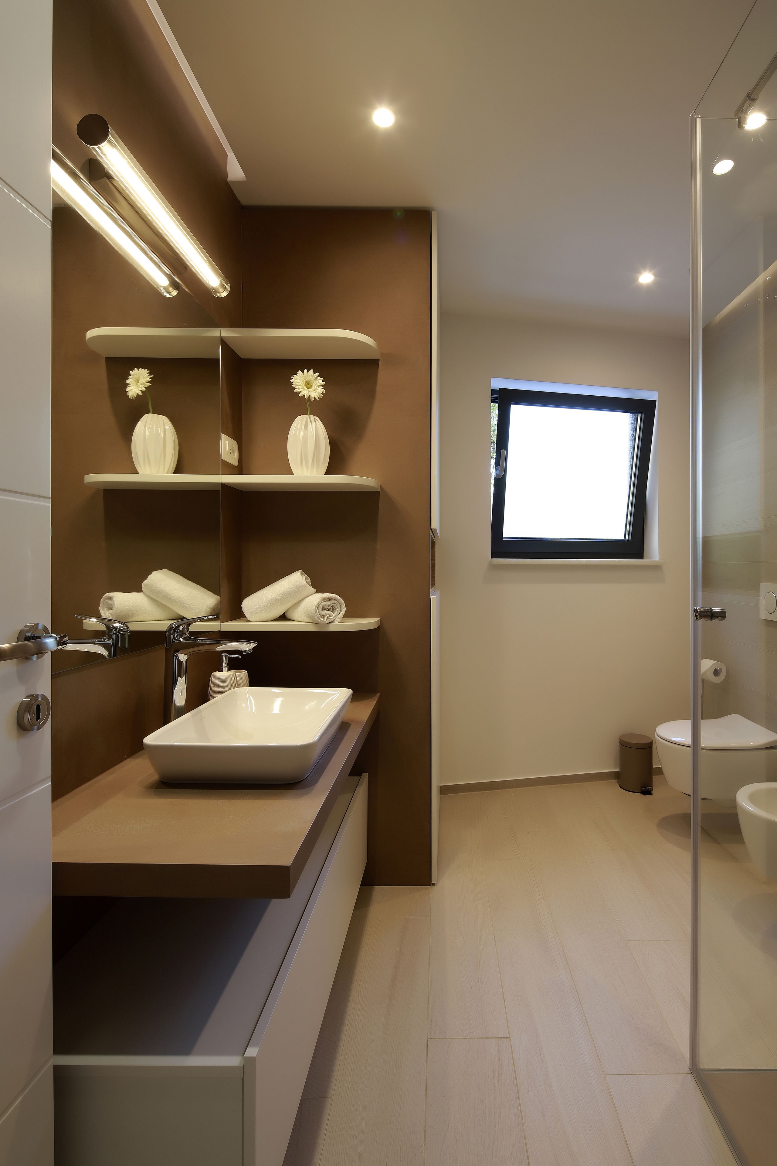 Un baño moderno con un espejo grande, un lavamanos blanco y un inodoro. Hay una ventana que proporciona luz natural y un estante con adornos.