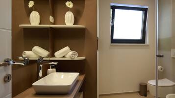 Un baño moderno con un espejo grande, un lavamanos blanco y un inodoro. Hay una ventana que proporciona luz natural y un estante con adornos.