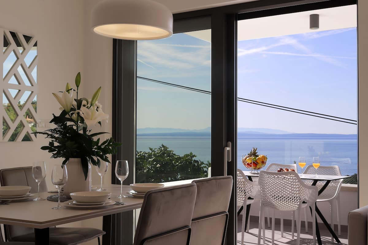 A dining area with a table set for two, a vase of flowers, and a view of the sea.