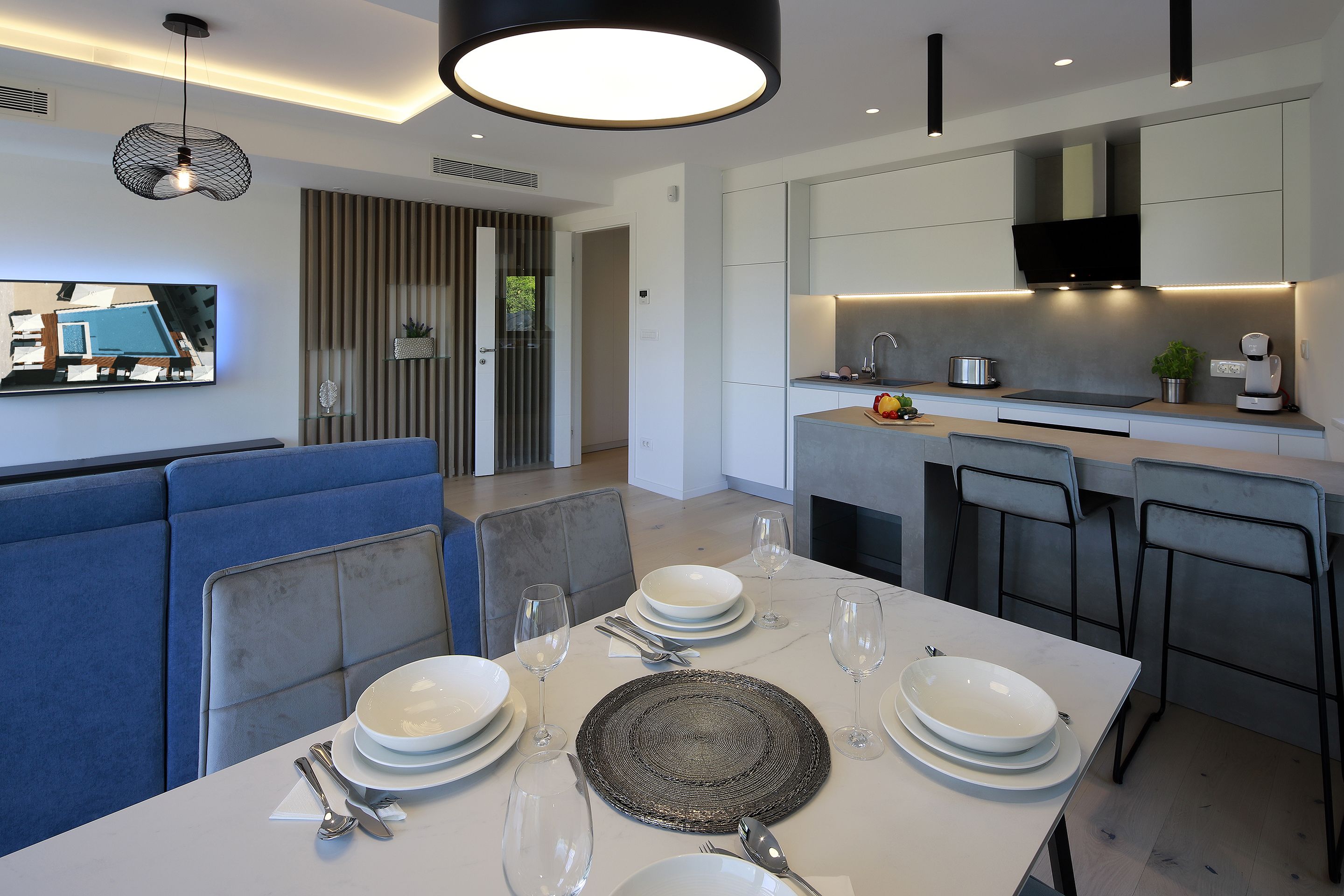 Un comedor moderno con una mesa blanca preparada para cuatro comensales, un sofá azul y una cocina con un televisor de pantalla plana sobre la encimera.