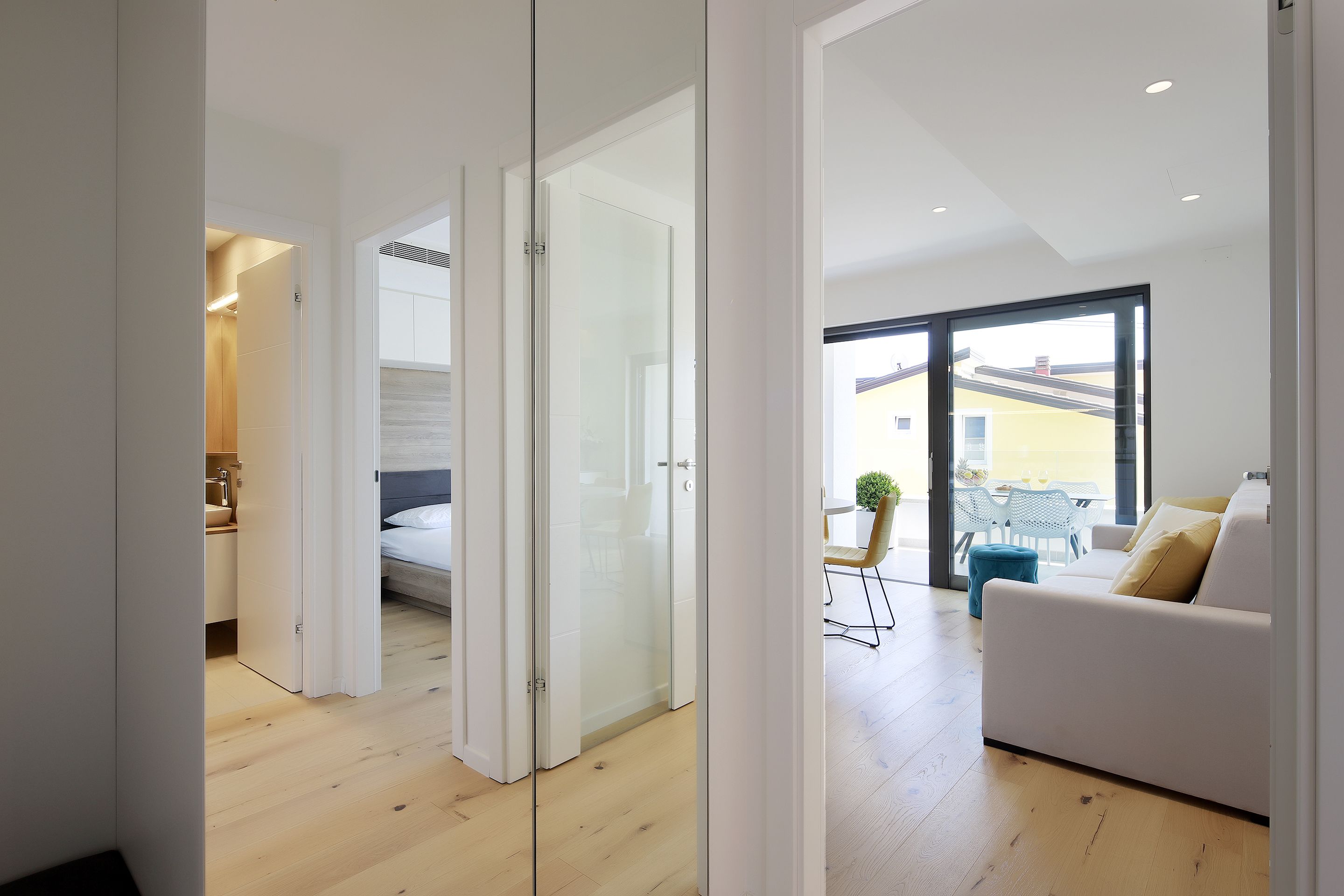 A modern interior with a wooden floor, a glass door leading to a bedroom, and a balcony with a white sofa and a table.