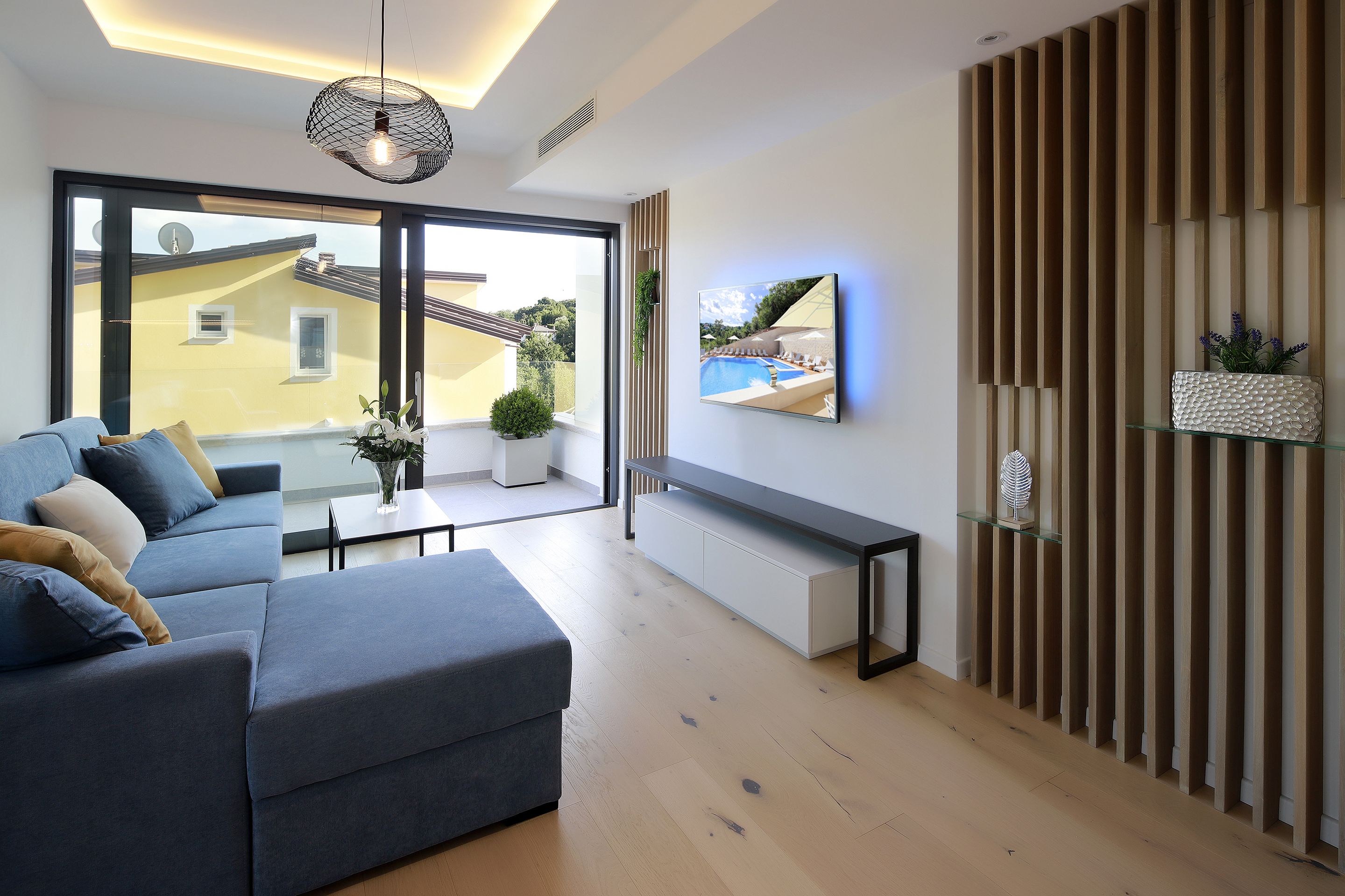 Un salon moderne avec un canapé gris, une table basse en verre et une grande fenêtre donnant sur un immeuble jaune et une piscine.
