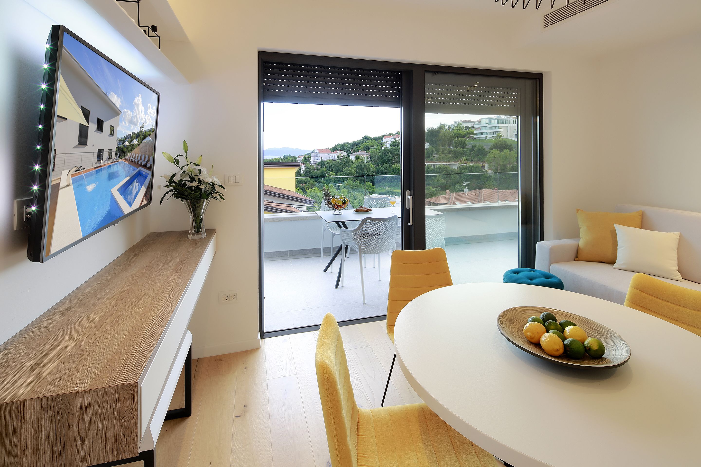 A modern living room with a dining area, a large window with a view of a residential area, and a balcony with outdoor furniture.