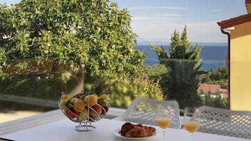 Et bord med en skål frugt, croissanter og glas appelsinjuice, med udsigt over et naturskønt hav- og grøntlandskab.