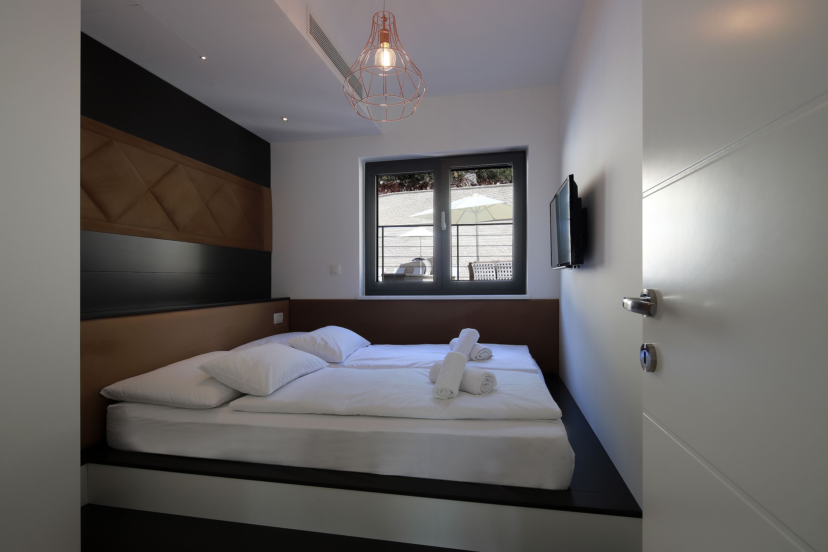 Une chambre moderne avec un grand lit, une tête de lit, une fenêtre avec des stores et un téléviseur fixé au mur.