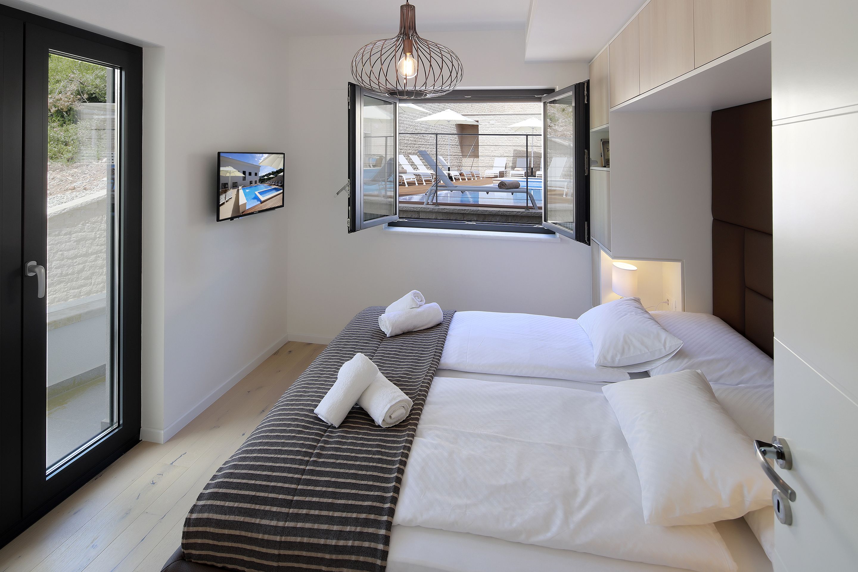 Una habitación de hotel moderna con un ventanal grande, una cama con sábanas blancas y una colcha a rayas, además de una lámpara colgante.