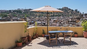 Une terrasse sur le toit, avec une table et des chaises sous un parasol, entourée de plantes en pot, d’où l’on peut admirer un paysage urbain avec des bâtiments et une colline en arrière-plan.