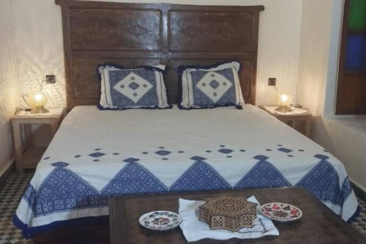 A bedroom with a wooden headboard, two bedside lamps, a bed with patterned bedding, a small table with a decorative box and plates, and a checkered floor.