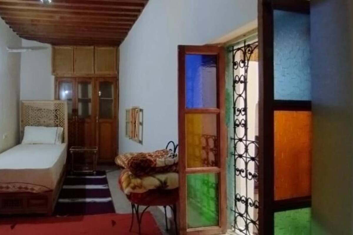 A compact room with a bed, a small table, and a chair. There is a door leading to another room and a window with a view.